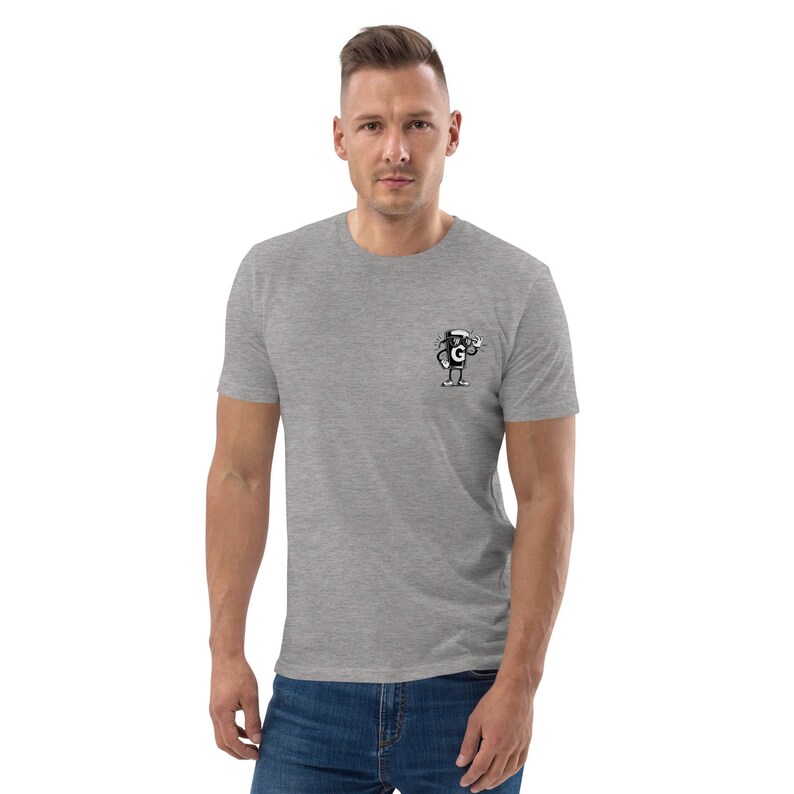Puede incluir: Una camiseta gris jaspeada con un peque&ntilde;o gr&aacute;fico en el pecho izquierdo. El gr&aacute;fico es un personaje de dibujos animados en blanco y negro que sostiene una taza de caf&eacute;. El personaje lleva un sombrero y gafas de sol. La camiseta es de manga corta y tiene cuello redondo.