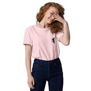 May include: A light pink t-shirt with a small black and white graphic on the chest. The graphic appears to be a cartoon character with a hat and a bow and arrow. The shirt is paired with dark blue jeans.