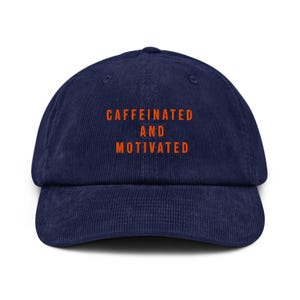 Puede incluir: Una gorra de béisbol de pana azul marino con el texto "CAFFEINATED AND MOTIVATED" bordado en naranja.