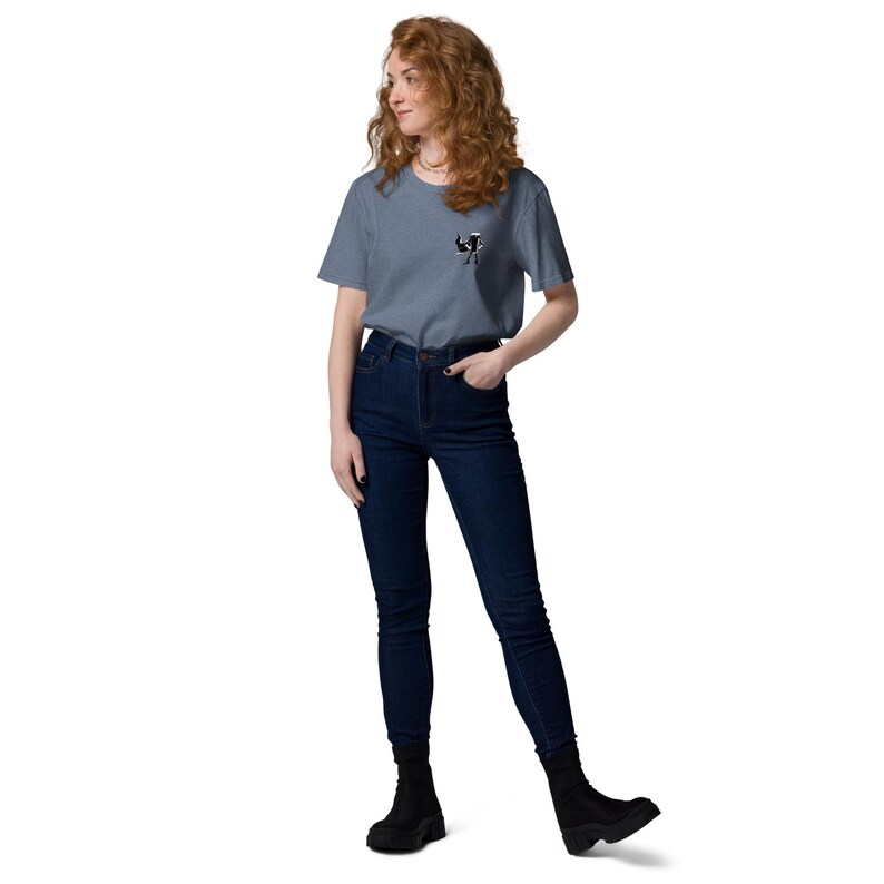 May include: A person wearing a blue t-shirt with a small black and white graphic on the chest, dark blue jeans and black boots. The t-shirt is plain, and the jeans are high-waisted.
