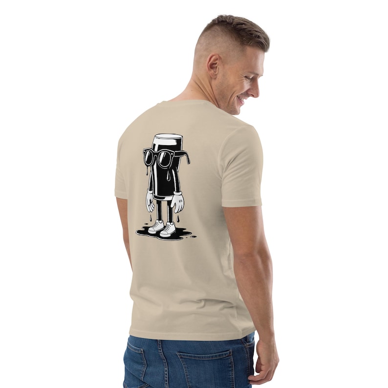 K&ouml;nnte beinhalten: Beiges T-Shirt mit einer Cartoon-Figur, die eine schwarze Fl&uuml;ssigkeit in einem Glas darstellt und eine Sonnenbrille, wei&szlig;e Handschuhe und Turnschuhe tr&auml;gt. Die Figur tropft auf eine schwarze Pf&uuml;tze. Das Shirt ist aus weichem Material.