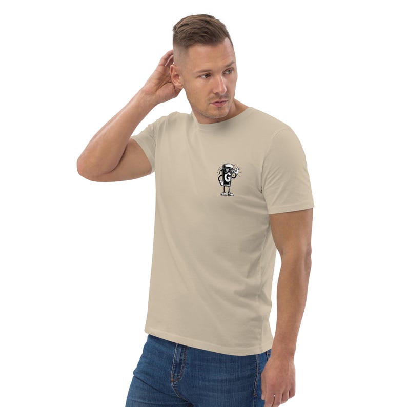 Puede incluir: Camiseta beige de manga corta con un peque&ntilde;o dibujo animado en blanco y negro en el pecho. El dibujo es un personaje estilizado con la letra "G". La camiseta es de material suave y de color neutro.