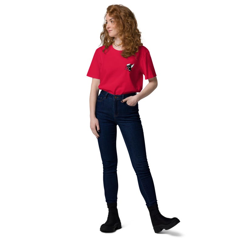 May include: A red t-shirt with a small graphic on the chest, paired with dark blue jeans and black ankle boots. The graphic appears to be a stylized design in black and white. The model is standing in front of a white background.