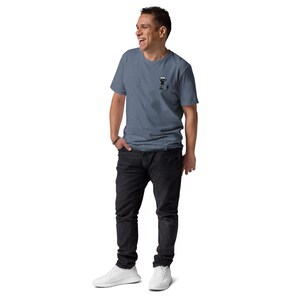 May include: A blue short-sleeve t-shirt with a small graphic on the left chest, paired with black jeans and white sneakers. The t-shirt has a crew neck and a subtle texture. The person is smiling and looking to the side.