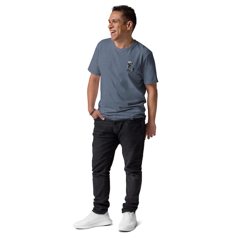 May include: A blue short-sleeve t-shirt with a small graphic on the left chest, paired with black jeans and white sneakers. The t-shirt has a crew neck and a subtle texture. The person is smiling and looking to the side.