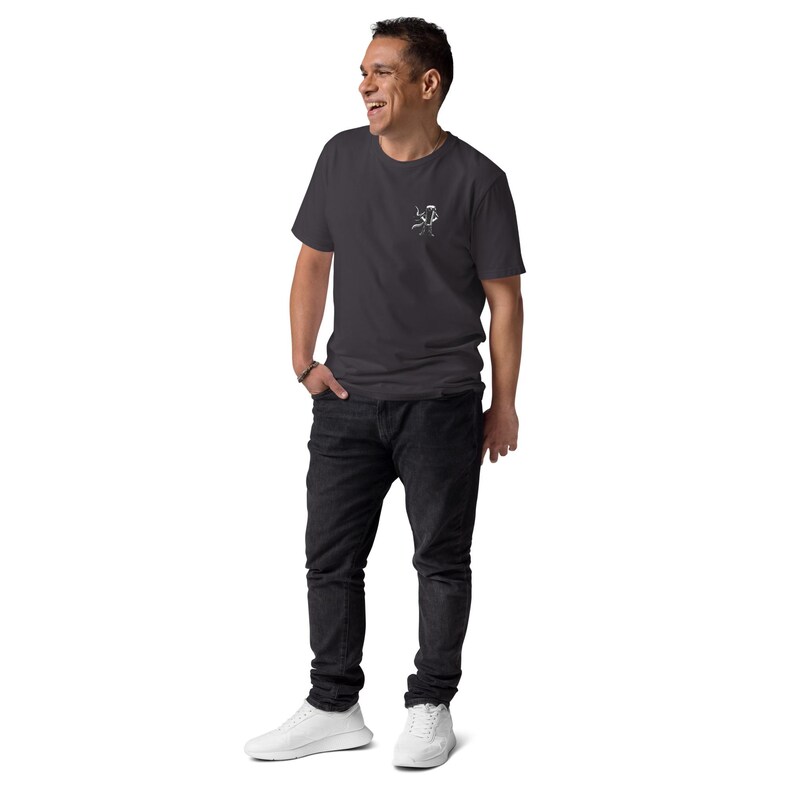 Puede incluir: Una camiseta gris oscuro con un peque&ntilde;o gr&aacute;fico blanco en el pecho, combinada con vaqueros negros y zapatillas blancas. La persona sonr&iacute;e y tiene una mano en el bolsillo.