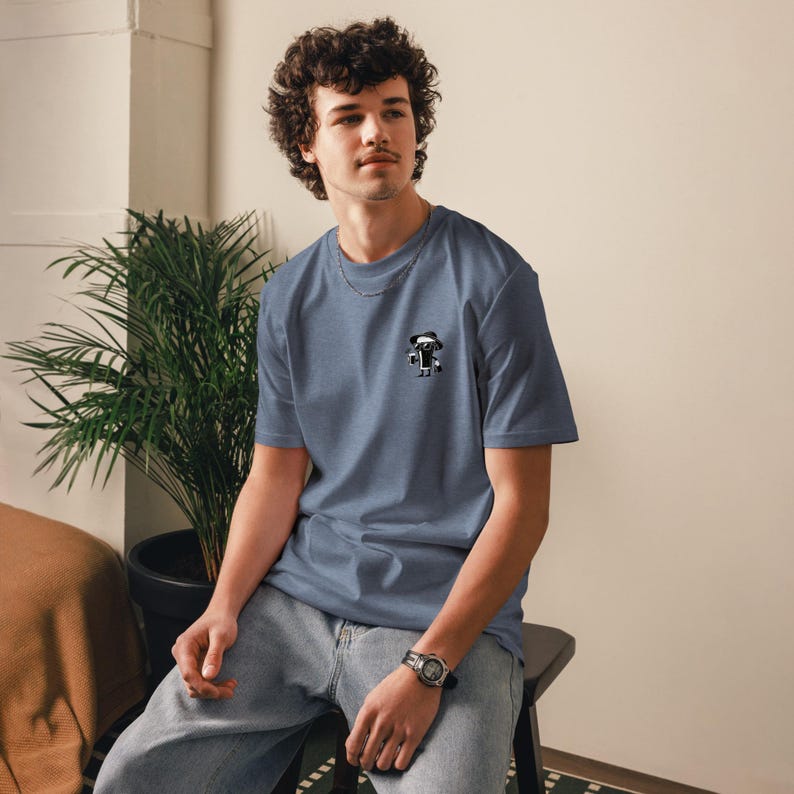 Puede incluir: Camiseta azul de manga corta con un peque&ntilde;o gr&aacute;fico en blanco y negro en el pecho. La persona lleva un collar de cadena plateada, un reloj y vaqueros azules. Una planta en maceta est&aacute; en el fondo.