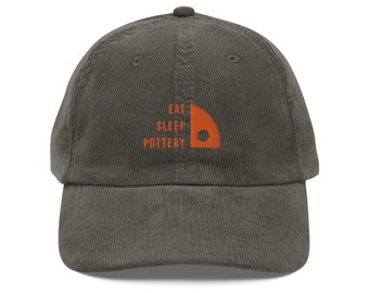 Może przedstawiać: Czapka bejsbolowa z ciemnozielonego, prążkowanego materiału welurowego z haftowanym pomarańczowym logo z napisem "Eat Sleep Pottery" i stylizowanym kształtem domu.