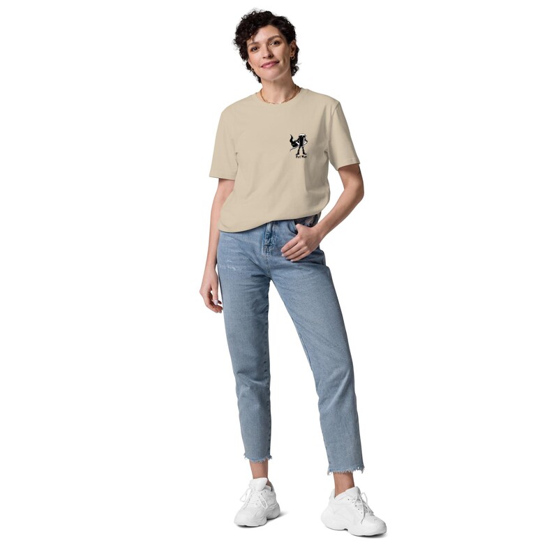 Puede incluir: Una persona con una camiseta beige con un peque&ntilde;o gr&aacute;fico negro en el pecho izquierdo, combinada con vaqueros azul claro y zapatillas blancas. La camiseta tiene cuello redondo y mangas cortas. Los vaqueros son rectos.