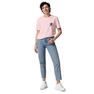 May include: A light pink t-shirt with a small black graphic on the chest, paired with light blue jeans and white sneakers. The t-shirt has short sleeves and a crew neck. The jeans are straight-legged with a raw hem.