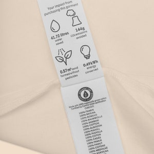 Może przedstawiać: Biała metka z czarnym tekstem i ikonami, kt&oacute;re szczeg&oacute;łowo opisują wpływ produktu na środowisko. Metka informuje, że zaoszczędzono 41,72 litr&oacute;w wody, uniknięto 1448 gram&oacute;w emisji CO2, 0,57 metra kwadratowego ziemi uprawiano bez pestycyd&oacute;w, a zaoszczędzono 0,49 kilowatogodziny energii. Metka informuje r&oacute;wnież, że produkt został wykonany ze 100% organicznie uprawianej bawełny i jest certyfikowany przez Control Union certification CU811271. Metka zawiera słowo "bawełna" w 12 r&oacute;żnych językach.