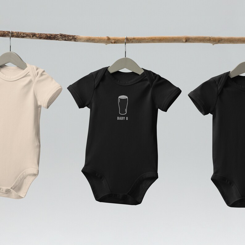 Może przedstawiać: Trzy body dla niemowląt wiszące na gałęzi. Body w środku jest czarne z białym obrysem szklanki piwa i tekstem "BABY G". Body po lewej stronie jest beżowe, a body po prawej stronie jest czarne.
