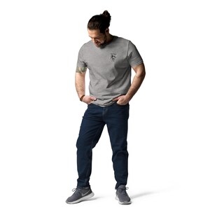 May include: A person wearing a grey t-shirt with a small graphic on the chest, dark blue jeans, and grey athletic shoes. The person is standing with their hands in their pockets.