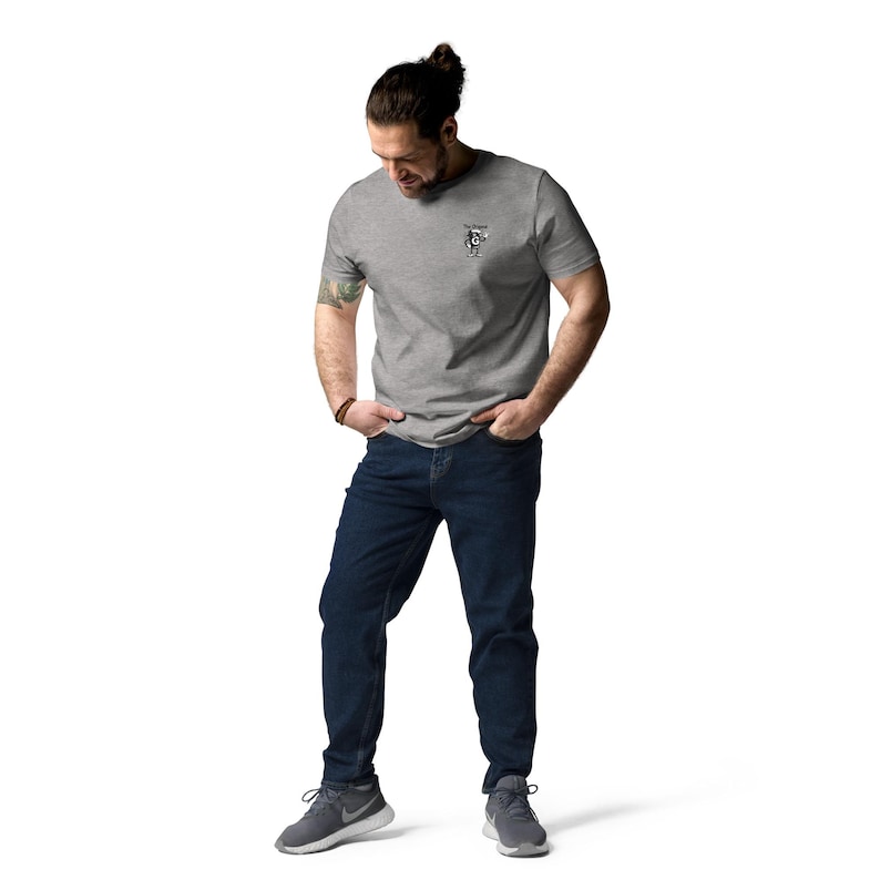 May include: A person wearing a grey t-shirt with a small graphic on the chest, dark blue jeans, and grey athletic shoes. The person is standing with their hands in their pockets.