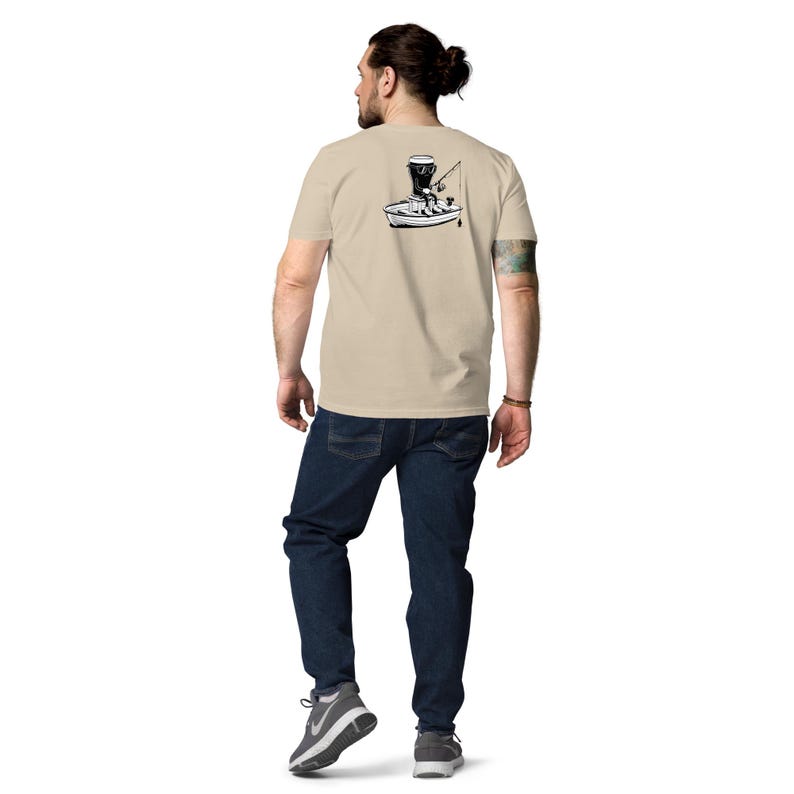 May include: Beige t-shirt with a black and white graphic of a person fishing from a boat. The person is wearing a hat and holding a fishing rod. The person is wearing blue jeans and gray sneakers.