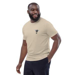 May include: Beige short-sleeve t-shirt with a small black and white graphic of a figure with wings on the chest. The shirt is made of a soft material and has a classic crew neck design. The model is wearing navy blue pants.