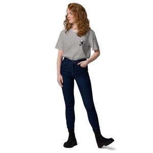 Puede incluir: Una camiseta gris claro con un peque&ntilde;o gr&aacute;fico en blanco y negro en el pecho izquierdo. La camiseta se combina con vaqueros de cintura alta azul oscuro y botines negros. La modelo est&aacute; de pie frente a un fondo blanco.