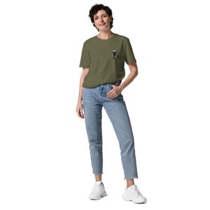 May include: An olive green t-shirt with a small graphic on the chest. The graphic appears to be a black and white illustration. The shirt is paired with light blue jeans and white sneakers. The model is standing in front of a white background.