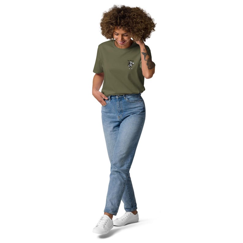 May include: An olive green t-shirt with a small graphic on the front. The shirt is worn with light blue jeans and white trainers. The outfit has a casual and relaxed style.