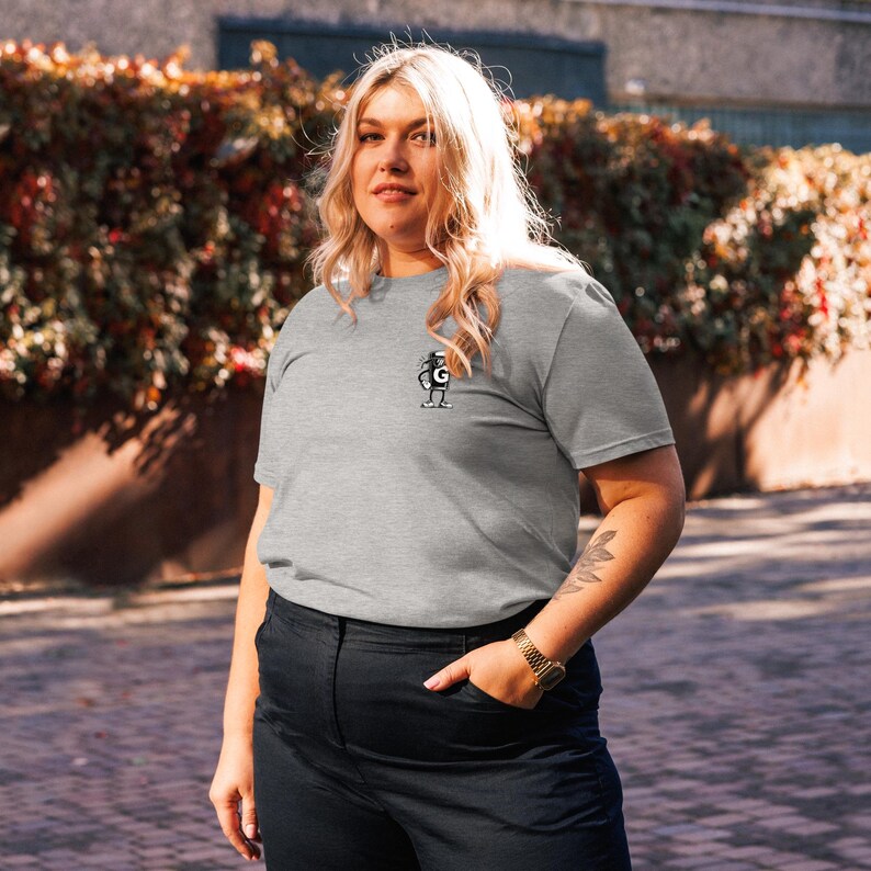 Puede incluir: Una camiseta gris con un peque&ntilde;o gr&aacute;fico en blanco y negro en el pecho. El gr&aacute;fico es una figura estilizada. La persona lleva pantalones negros y un reloj dorado.