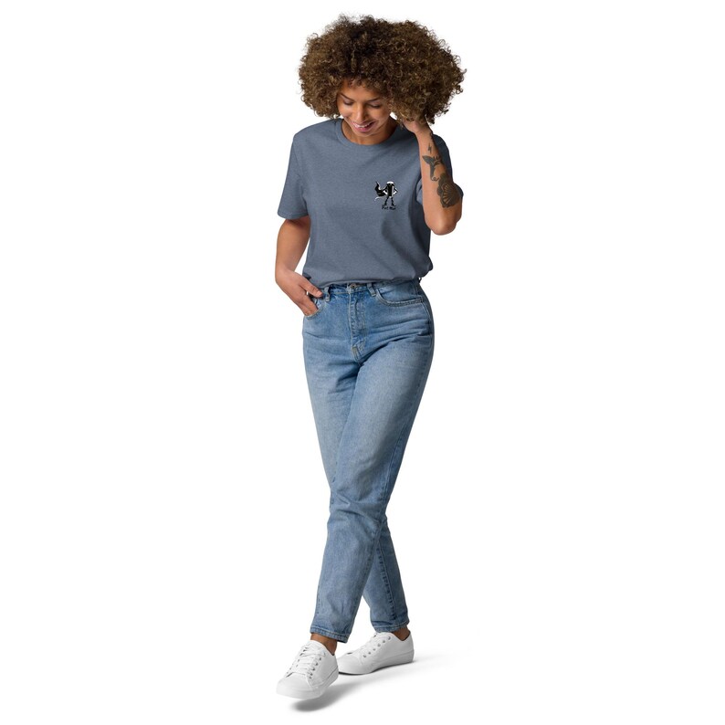 Puede incluir: Una camiseta azul con un peque&ntilde;o gr&aacute;fico en blanco y negro. Se combina con vaqueros azul claro y zapatillas blancas. La persona est&aacute; de pie frente a un fondo blanco.