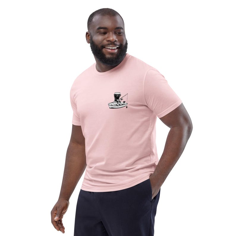 May include: A light pink t-shirt featuring a small black and white graphic of a person fishing from a boat. The t-shirt is short-sleeved and has a crew neck. The person is wearing dark blue pants.