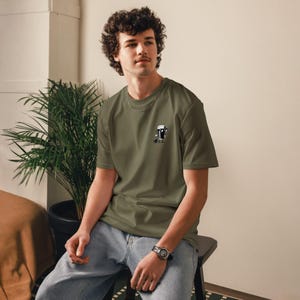 Puede incluir: Camiseta verde oliva con un peque&ntilde;o gr&aacute;fico en blanco y negro en el pecho. El gr&aacute;fico incluye texto japon&eacute;s. La camiseta es de cuello redondo y se usa con jeans azules. La persona lleva un reloj.