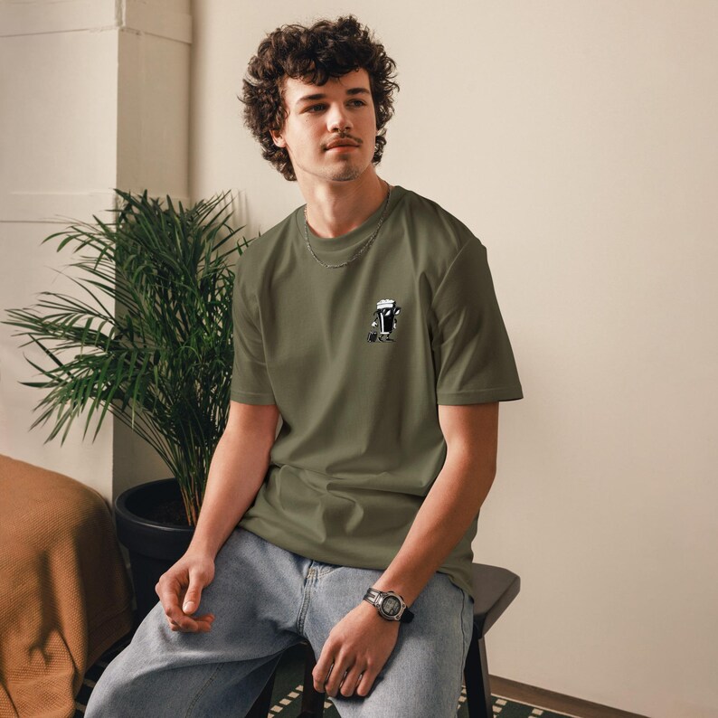 Puede incluir: Camiseta verde oliva con un peque&ntilde;o gr&aacute;fico en blanco y negro en el pecho. El gr&aacute;fico incluye texto japon&eacute;s. La camiseta es de cuello redondo y se usa con jeans azules. La persona lleva un reloj.