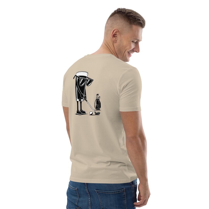 May include: Beige t-shirt with a black and white graphic of a cartoon beer mug playing golf. The beer mug wears sunglasses and is positioned to hit a golf ball. A golf bag and club are also in the graphic.