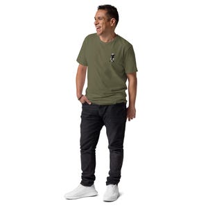 K&ouml;nnte beinhalten: Ein olivgr&uuml;nes T-Shirt mit einer kleinen Grafik auf der linken Brust. Die Person tr&auml;gt eine schwarze Jeans und wei&szlig;e Turnschuhe. Die Person l&auml;chelt und blickt nach links.
