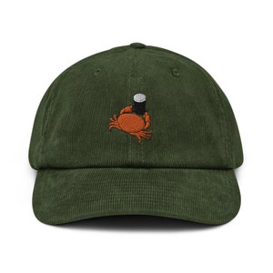 Può includere: Un berretto da baseball in velluto a coste verde con un granchio arancione ricamato che tiene una lattina nera e bianca.