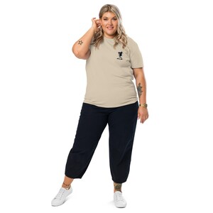 K&ouml;nnte beinhalten: Beiges T-Shirt mit kleinem schwarzem Aufdruck auf der Brust, kombiniert mit dunkelblauen, cropped Hosen und wei&szlig;en Turnschuhen. Die Person tr&auml;gt eine goldene Uhr.