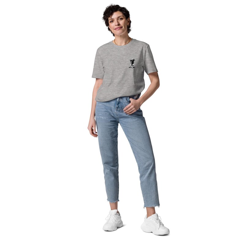 K&ouml;nnte beinhalten: Eine Person tr&auml;gt ein hellgraues T-Shirt mit einer kleinen schwarz-wei&szlig;en Grafik auf der Brust, kombiniert mit hellblauen Jeans und wei&szlig;en Turnschuhen. Das T-Shirt ist kurz&auml;rmlig und die Jeans sind cropped. Der Stil ist l&auml;ssig.