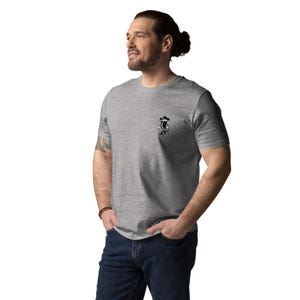 May include: A heather gray t-shirt featuring a small black and white cartoon cowboy graphic on the chest. The shirt has short sleeves and a crew neck. The person is wearing blue jeans.