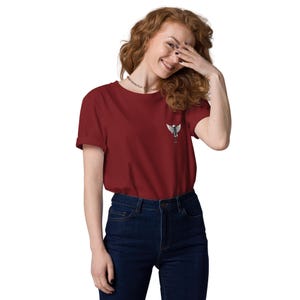 May include: A maroon t-shirt with a small white and gray winged emblem on the chest. The shirt has short sleeves and is paired with dark blue jeans. The person is smiling and covering their face with their hand.