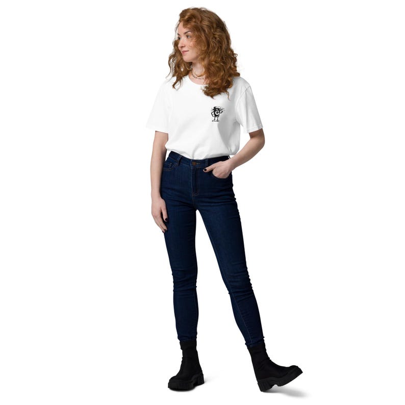 Puede incluir: Una camiseta blanca con un peque&ntilde;o gr&aacute;fico en blanco y negro. La persona lleva vaqueros azul oscuro y botas negras. La persona est&aacute; de pie frente a un fondo blanco.