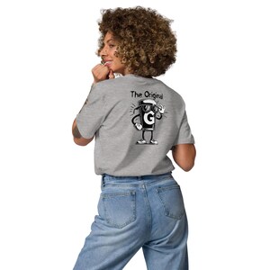 May include: A light grey t-shirt with a cartoon coffee cup graphic on the back, wearing sunglasses and giving a thumbs-up. The text "The Original" is above the graphic. The person is wearing blue jeans.