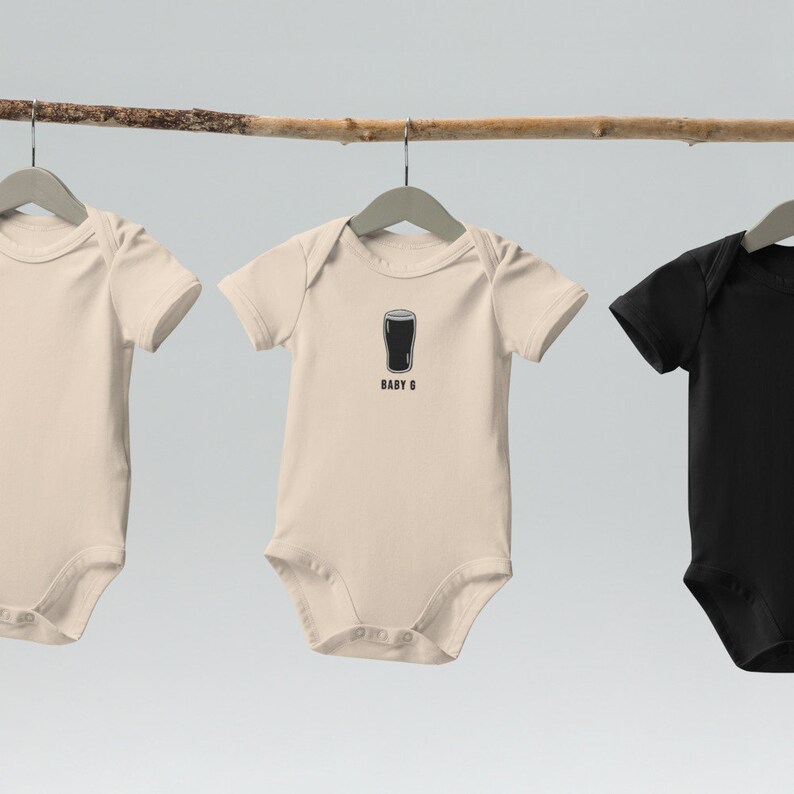 Może przedstawiać: Dwa body dla niemowląt wiszące na drewnianym gałęzi. Jeden body jest beżowy z czarnym nadrukiem szklanki piwa i tekstem "BABY G". Drugi body jest czarny.