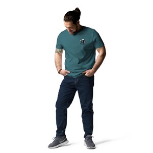 May include: Teal short-sleeve t-shirt with a small graphic on the left chest. The graphic appears to be a cartoon character. The person is wearing dark blue jeans and gray athletic shoes.