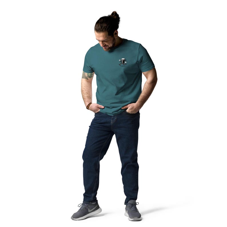 May include: Teal short-sleeve t-shirt with a small graphic on the left chest. The graphic appears to be a cartoon character. The person is wearing dark blue jeans and gray athletic shoes.