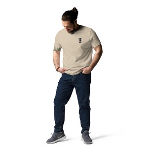 May include: A beige t-shirt with a small black graphic on the chest. The t-shirt is paired with dark blue jeans and gray athletic shoes. The person is standing with their hands in their pockets.