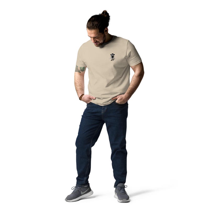 May include: A beige t-shirt with a small black graphic on the chest. The t-shirt is paired with dark blue jeans and gray athletic shoes. The person is standing with their hands in their pockets.