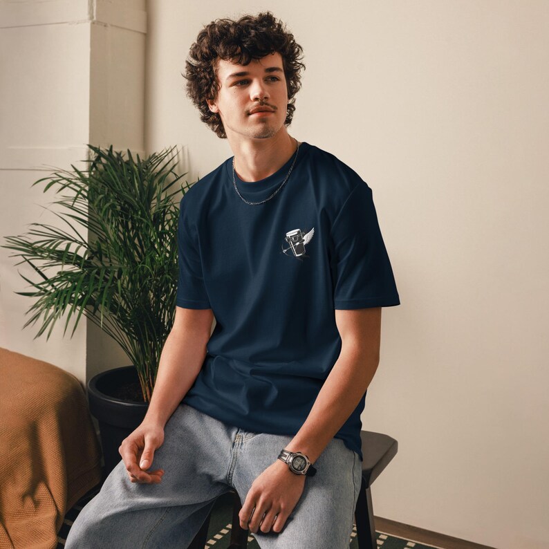May include: Navy blue t-shirt with a small white graphic of a coffee cup with wings on the left chest. The shirt is a short-sleeved, crew neck style. The person is wearing a silver chain necklace and a watch.