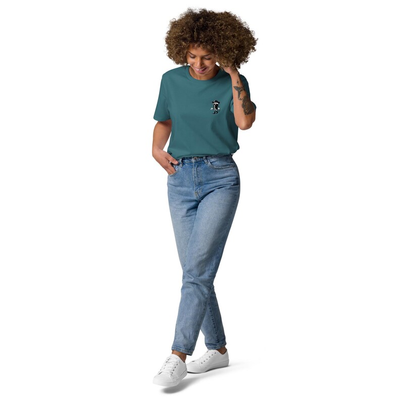 May include: Teal-colored t-shirt with a small graphic on the chest, paired with light blue jeans and white sneakers. The t-shirt features a small black and white graphic. The model is wearing a casual outfit.