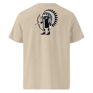 May include: Beige t-shirt featuring a black and white cartoon design on the back. The design depicts a cartoon character wearing sunglasses and a Native American headdress, holding a bow and arrow. The shirt is made of a soft, neutral-colored fabric.