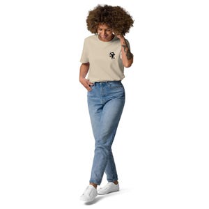 Puede incluir: Una mujer con una camiseta beige con un peque&ntilde;o gr&aacute;fico negro en el pecho, combinada con vaqueros azules claros y zapatillas blancas. La camiseta tiene cuello redondo y mangas cortas. Los vaqueros son de lavado claro.