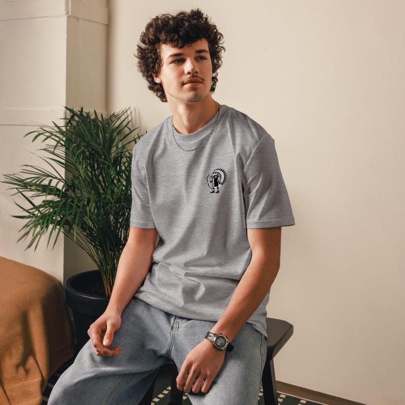 May include: A heather gray t-shirt with a small black and white graphic on the chest. The graphic depicts a stylized Native American figure. The shirt is short-sleeved and has a crew neck.