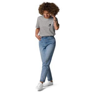 Puede incluir: Una camiseta gris claro con un peque&ntilde;o gr&aacute;fico en blanco y negro en el pecho. La camiseta se combina con vaqueros azul claro y zapatillas blancas. La persona est&aacute; de pie frente a un fondo blanco.