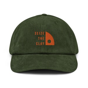 Op de afbeelding: Een groene corduroy baseballpet met een oranje geborduurd logo met de tekst "Seize the Clay" en een kleine oranje cirkel.