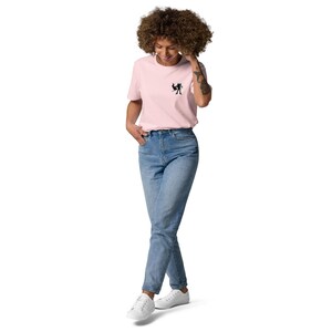 May include: A pale pink t-shirt with a small black graphic on the chest, worn with blue jeans and white trainers. The t-shirt has short sleeves and a crew neck. The jeans are a light wash, and the trainers are low-top.